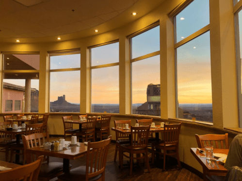 A cozy restaurant with wooden tables and chairs, featuring large windows showcasing a colorful sunset view.