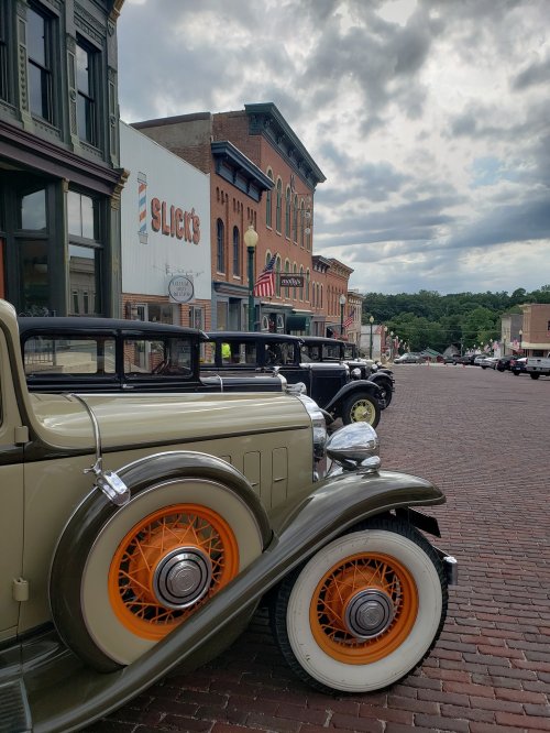 Vintage cars parked on a cobblestone street, with historic buildings and a cloudy sky in the background.