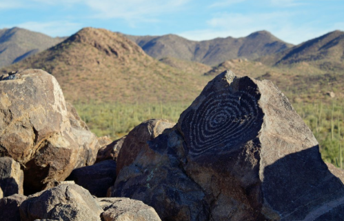 Petroglyph of a spiral design on a rock, with mountains and desert vegetation in the background.