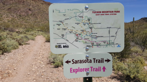 Trail sign at Tucson Mountain Park showing map and directions to Sarasota Trail and Explorer Trail. Desert landscape in background.