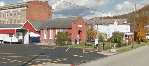 A red brick museum building with a sign, surrounded by trees and parked cars on a sunny day.