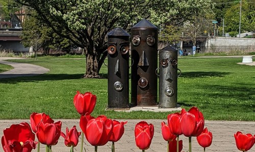 Three whimsical metal sculptures with faces stand in a park, surrounded by vibrant red tulips.
