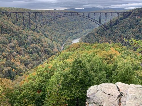A scenic view of a valley with a large bridge spanning across, surrounded by colorful autumn foliage and cloudy skies.
