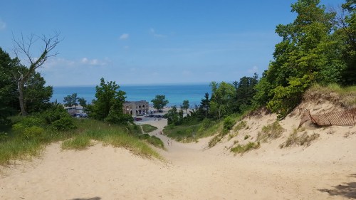 A sandy dune leads down to a blue lake, surrounded by green trees and a clear sky. People walk in the distance.