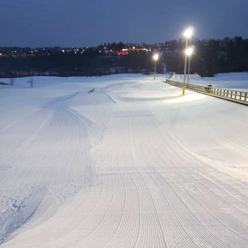 A snowy landscape at dusk, featuring groomed ski trails and illuminated by bright lights in the background.