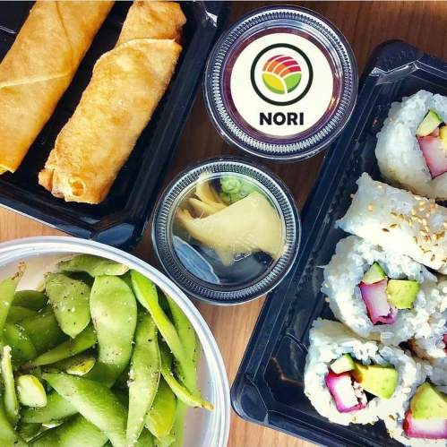 A variety of sushi rolls, spring rolls, edamame, and pickled ginger in takeout containers. Nori logo visible.
