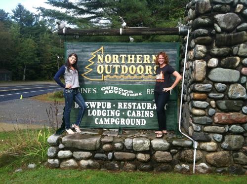 Two women pose beside a stone sign for Northern Outdoors, featuring outdoor adventure and lodging information.