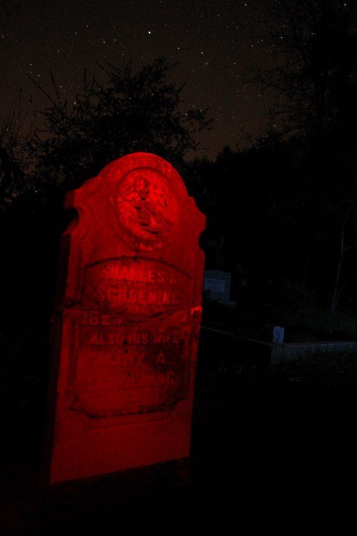 A red-lit gravestone under a starry night sky, featuring the name "Charles Schrenne" and a decorative emblem.