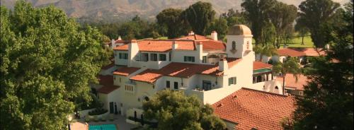 Aerial view of a large, elegant white villa with red tile roofs, surrounded by trees and mountains in the background.