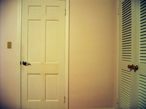 A simple room with a white door on the left and a closed louvered closet door on the right against a pink wall.