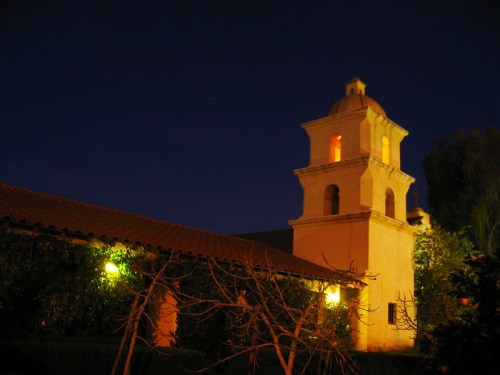 A historic building with a bell tower illuminated at night, surrounded by trees and a dark blue sky.