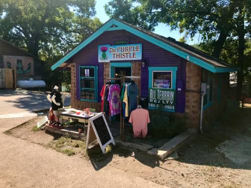 A colorful storefront called "The Purple Thistle" with clothing displayed outside and a sign for All Terrain Realty.