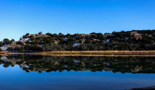 A serene landscape featuring a snowy hillside reflected in calm water under a clear blue sky.