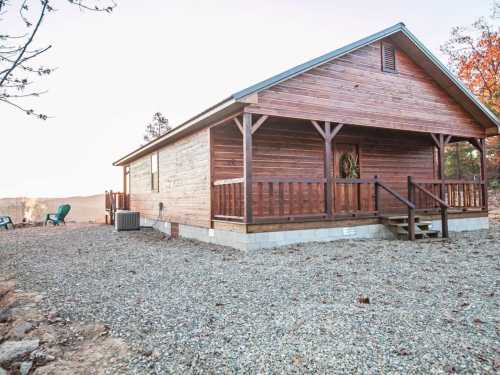 A wooden cabin with a porch, surrounded by gravel and trees, set against a scenic landscape.