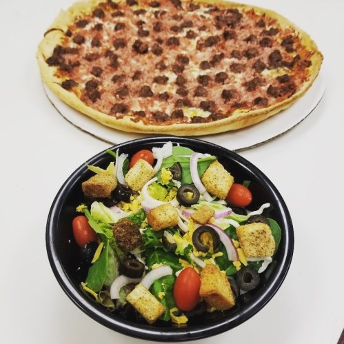 A bowl of fresh salad with tomatoes, olives, and croutons, alongside a large meat-topped pizza.