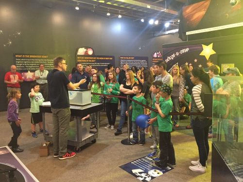 A group of people, including children, watch a science demonstration in a museum exhibit about stars.