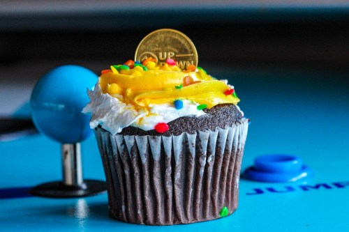 A chocolate cupcake topped with yellow frosting, sprinkles, and a gold coin, set against a blue background.