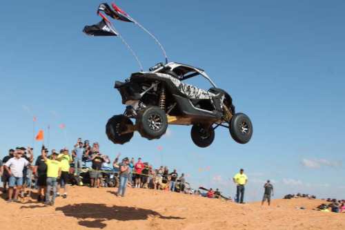 A dune buggy jumps over a crowd at a sandy event, with flags waving in the background and spectators watching.