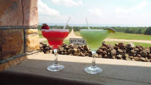Two colorful cocktails, one red and one green, sit on a stone ledge with a scenic view in the background.
