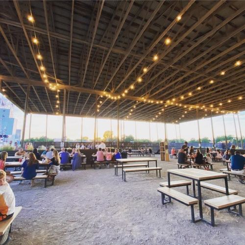 A spacious outdoor venue with string lights, filled with people seated at picnic tables during sunset.