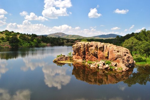 A serene lake scene with a rocky outcrop, surrounded by lush greenery and blue skies with fluffy clouds reflected in the water.
