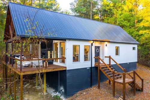 A modern cabin with a metal roof, wooden deck, and stairs, surrounded by trees in a natural setting.