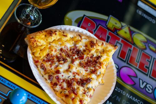 A slice of cheesy bacon pizza on a paper plate, next to a glass of beer and an arcade game screen in the background.