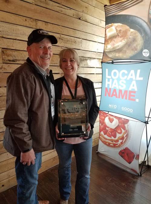 A man and woman pose together, holding an award, in front of a wooden wall and a promotional banner.