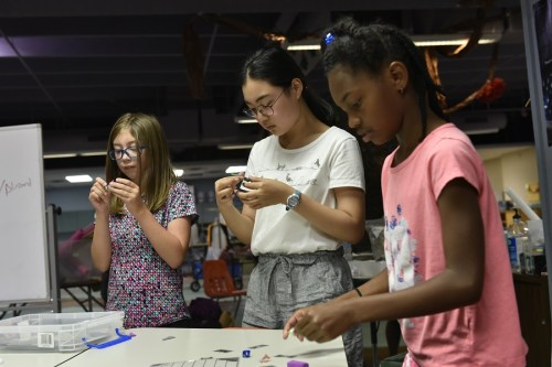 Three young girls are focused on crafting with small materials at a table in a creative workspace.