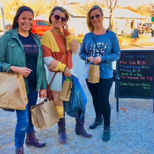 Three women stand smiling with takeout bags, in front of a chalkboard menu at an outdoor food market.