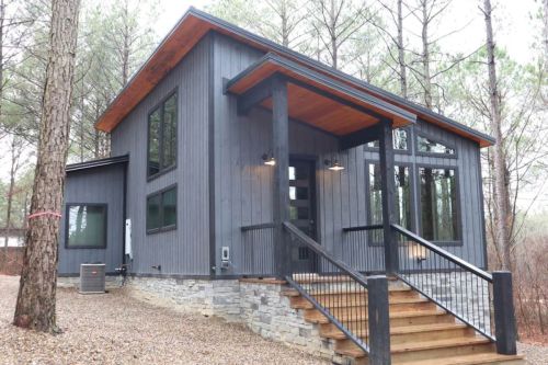 Modern gray house with large windows, wooden accents, and a stone foundation, surrounded by trees and gravel pathway.