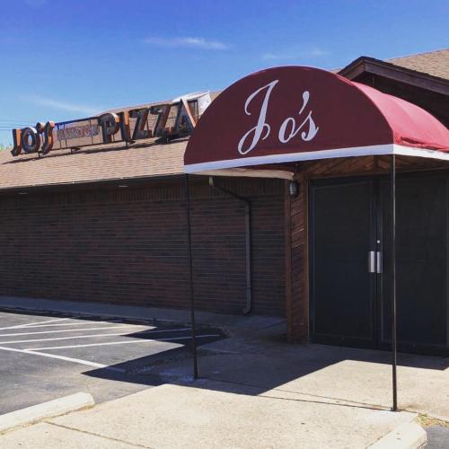 A red awning with "Jo's" above the entrance of a brick building, featuring a pizza sign in the background.