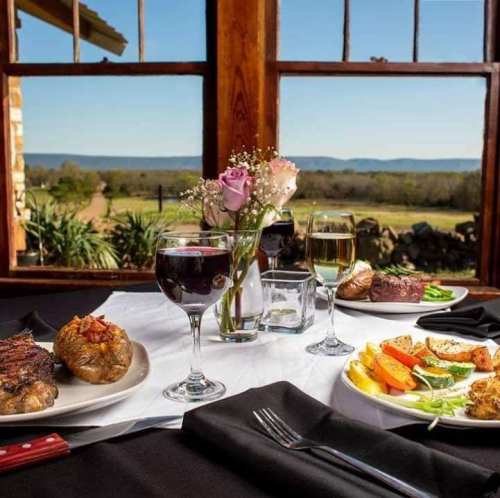 A beautifully set table with two plates of gourmet food, wine glasses, and a vase of flowers, overlooking a scenic view.