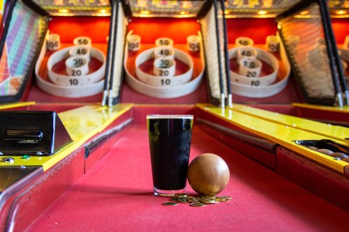 A pint of dark beer next to a wooden ball and coins, with a skee-ball game in the background.