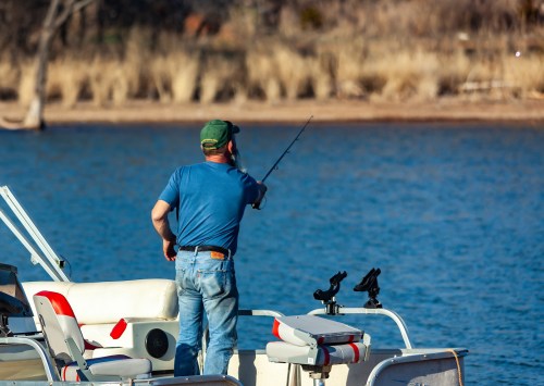 A man in a blue shirt and green cap fishing from a boat on a calm lake surrounded by dry grass.