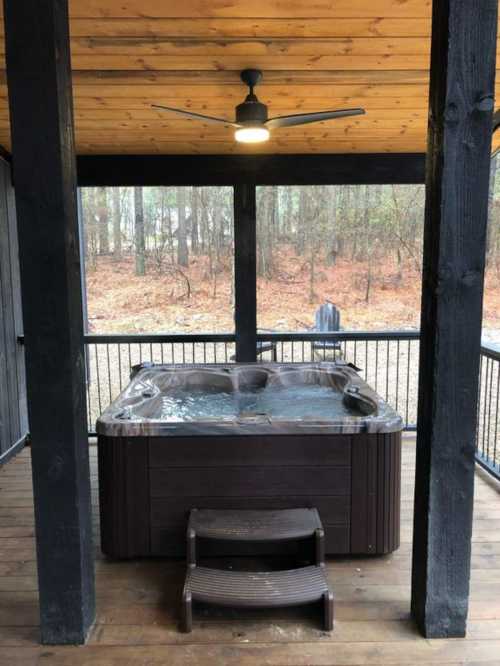 A wooden deck with a hot tub under a covered area, surrounded by trees and a chair in the background.