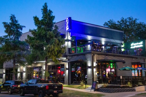 A lively restaurant with outdoor seating, illuminated at night, surrounded by trees and parked cars.