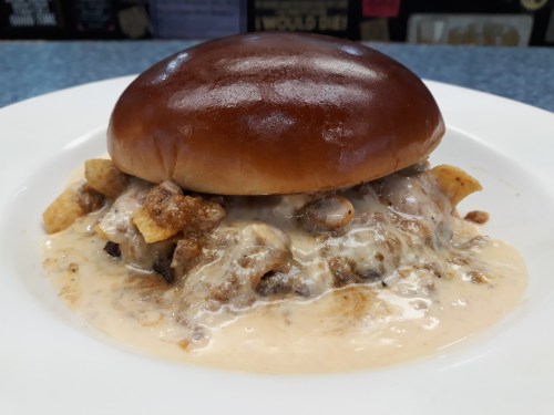 A close-up of a burger with a glossy bun, filled with creamy sauce and crunchy toppings on a white plate.