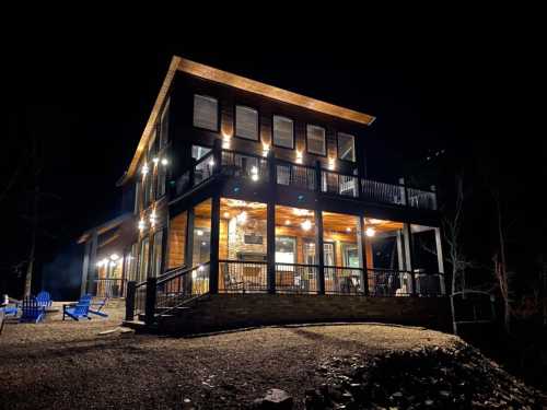 A modern cabin at night, illuminated with warm lights, surrounded by trees and featuring a deck and outdoor seating.