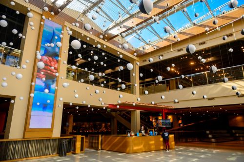 Bright interior of a modern building with hanging spheres, large screens, and a reception area. Natural light floods in.