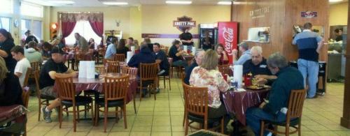 A busy diner with patrons at tables, a counter in the background, and a Coca-Cola machine visible.