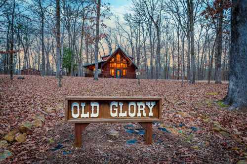 A wooden sign reading "OLD GLORY" in front of a rustic cabin surrounded by trees and fallen leaves at dusk.
