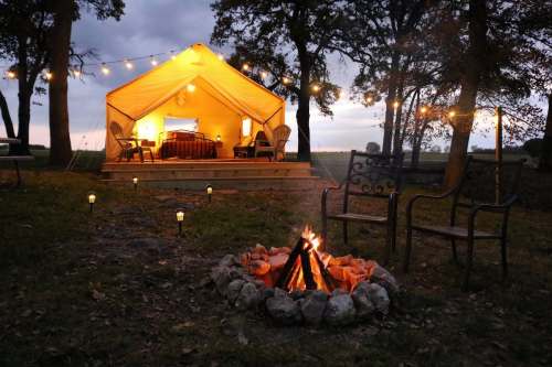 A cozy tent with string lights, a fire pit, and chairs, set in a serene outdoor environment at dusk.