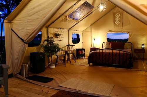 Cozy glamping tent interior with a bed, chairs, lanterns, and decorative plants, set in a serene outdoor environment.