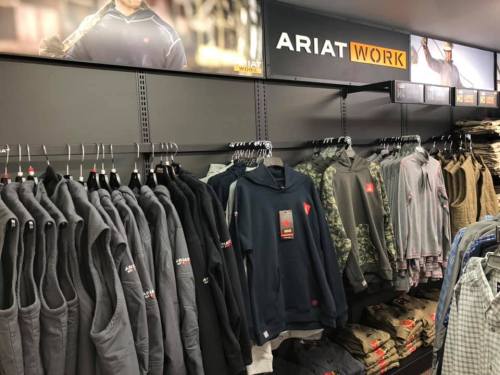 A display of Ariat Work clothing, featuring hoodies and vests, organized on racks in a retail store.