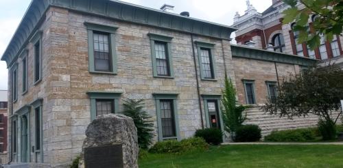 Historic stone building with green trim, surrounded by grass and trees, featuring a plaque in front.