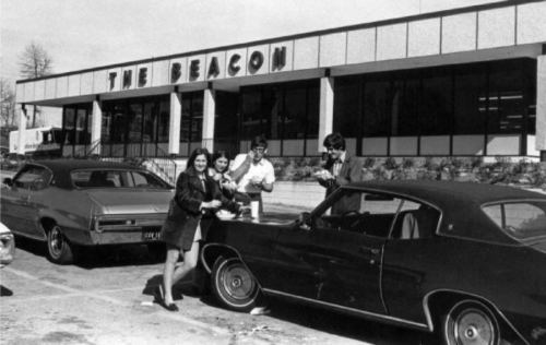 A group of five people stands by cars outside a building labeled "The Beacon," in a vintage black-and-white photo.