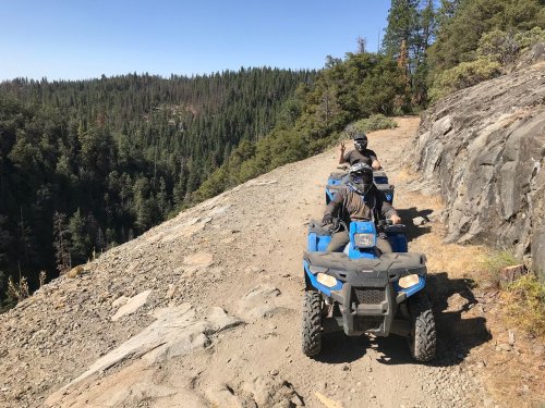 Two people riding blue ATVs along a rocky trail with a forested landscape in the background.