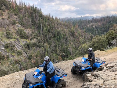 Two people riding blue ATVs on a rocky trail with a forested mountain landscape in the background.