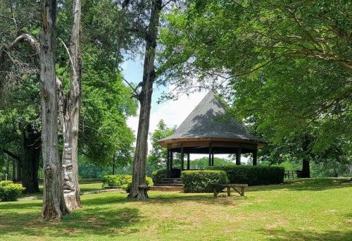 A gazebo surrounded by lush greenery and trees in a sunny park setting.